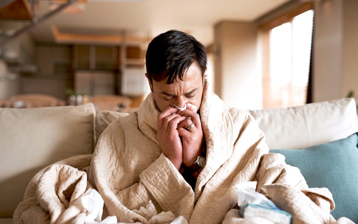 Cropped shot of a young man suffering with a cold while sitting wrapped in a blanket on the sofa at home.