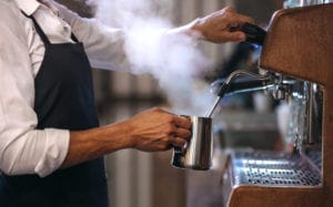Coffee shop worker preparing coffee on steam espresso coffee machine. Cropped shot of man working in coffee shop wearing an apron.