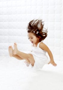 A happy young girl in white dress having fun jumping on mattress.