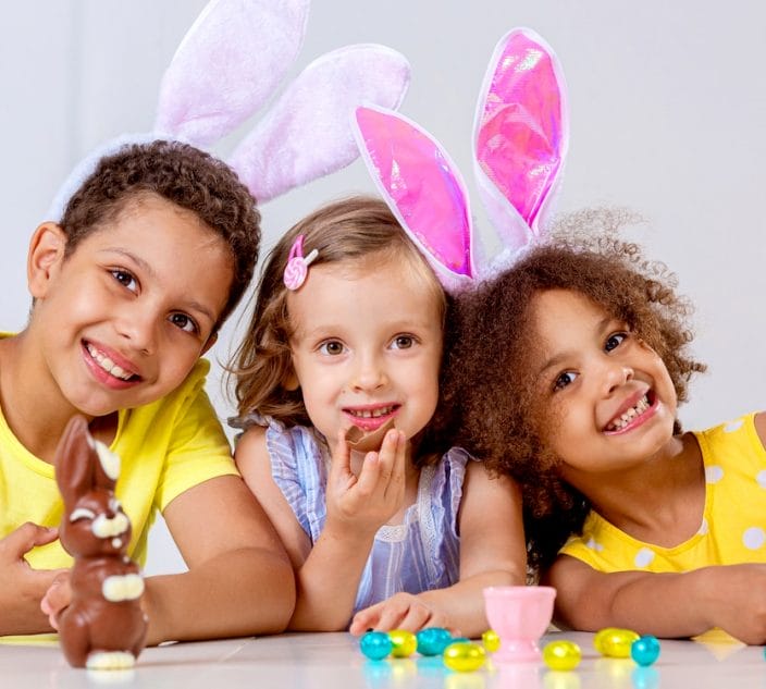 Three children in bright clothes and bunny ears eating Easter chocolate eggs sitting at the table at home