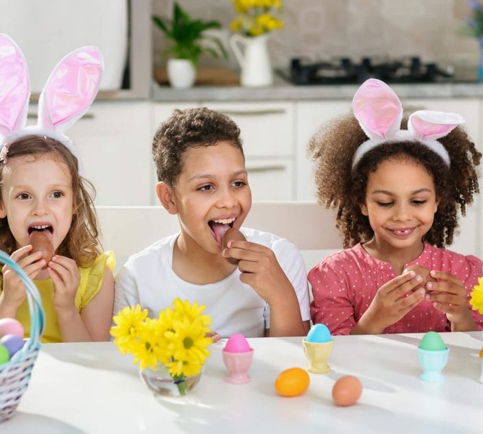three children in bright clothes and bunny ears are eating Easter chocolate eggs at home after the Easter egg Hunt
